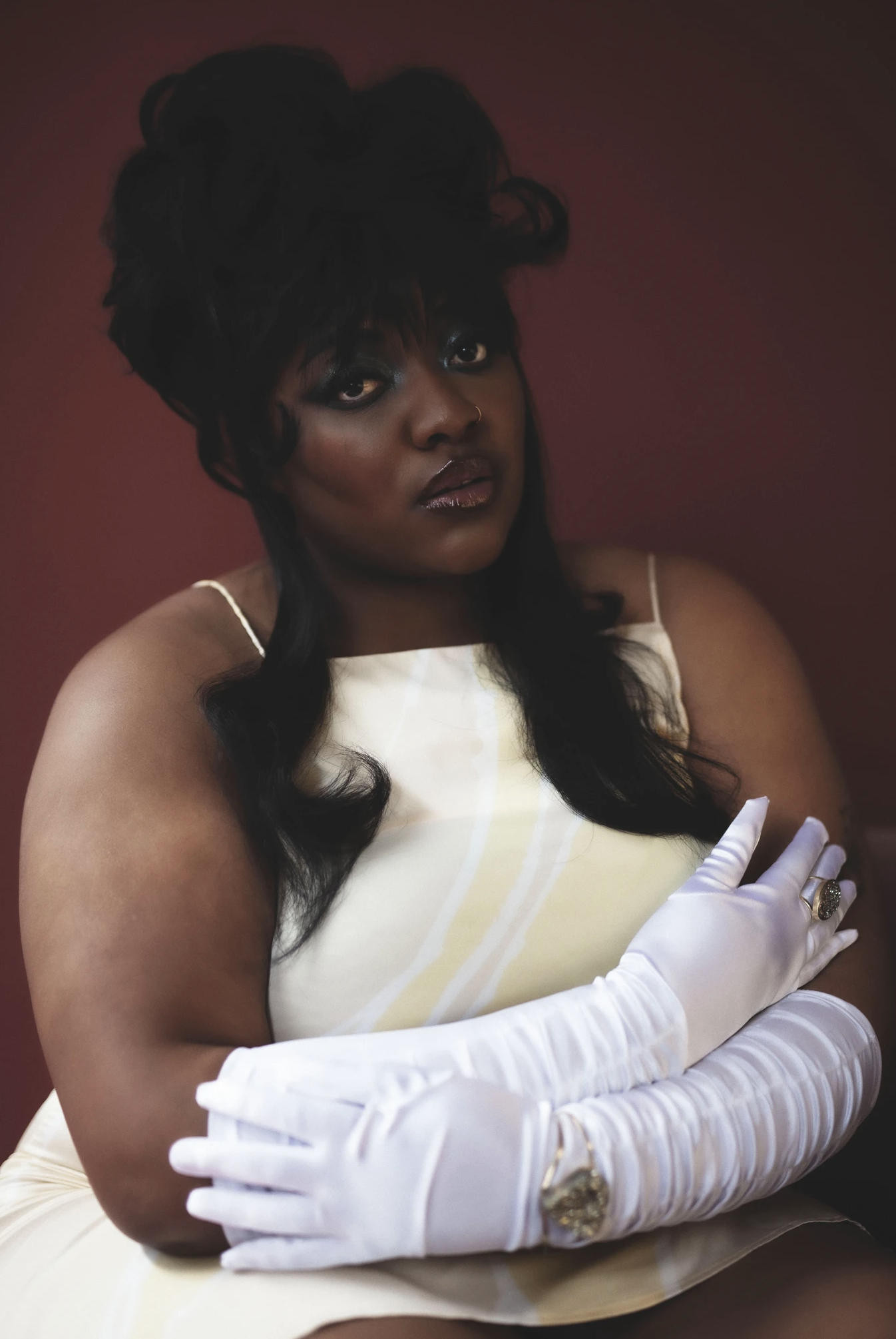 Portrait of a black woman with arms crossed wearing white gloves and a light yellow dress against a maroon background.