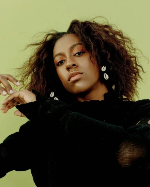 Portrait of a young adult woman with curly brown hair and neutral expression against a light green background, wearing shell earrings.