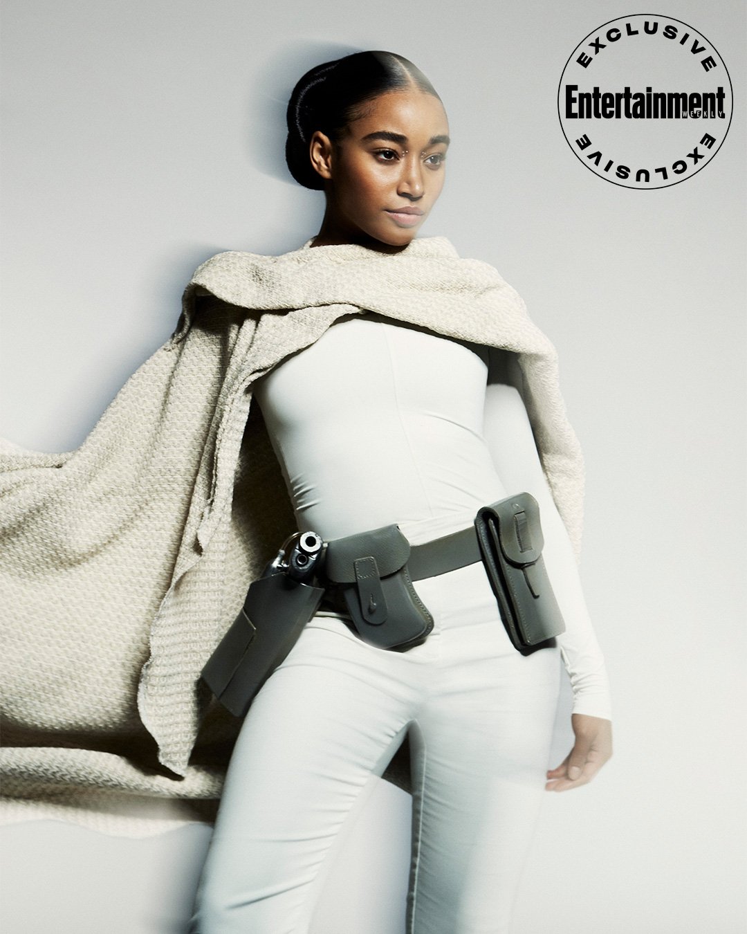Woman in all-white outfit accessorized with a utility belt holding a handgun, standing in front of a gradient grey backdrop. 