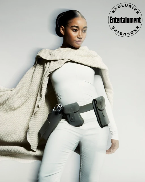Woman in all-white outfit accessorized with a utility belt holding a handgun, standing in front of a gradient grey backdrop. 