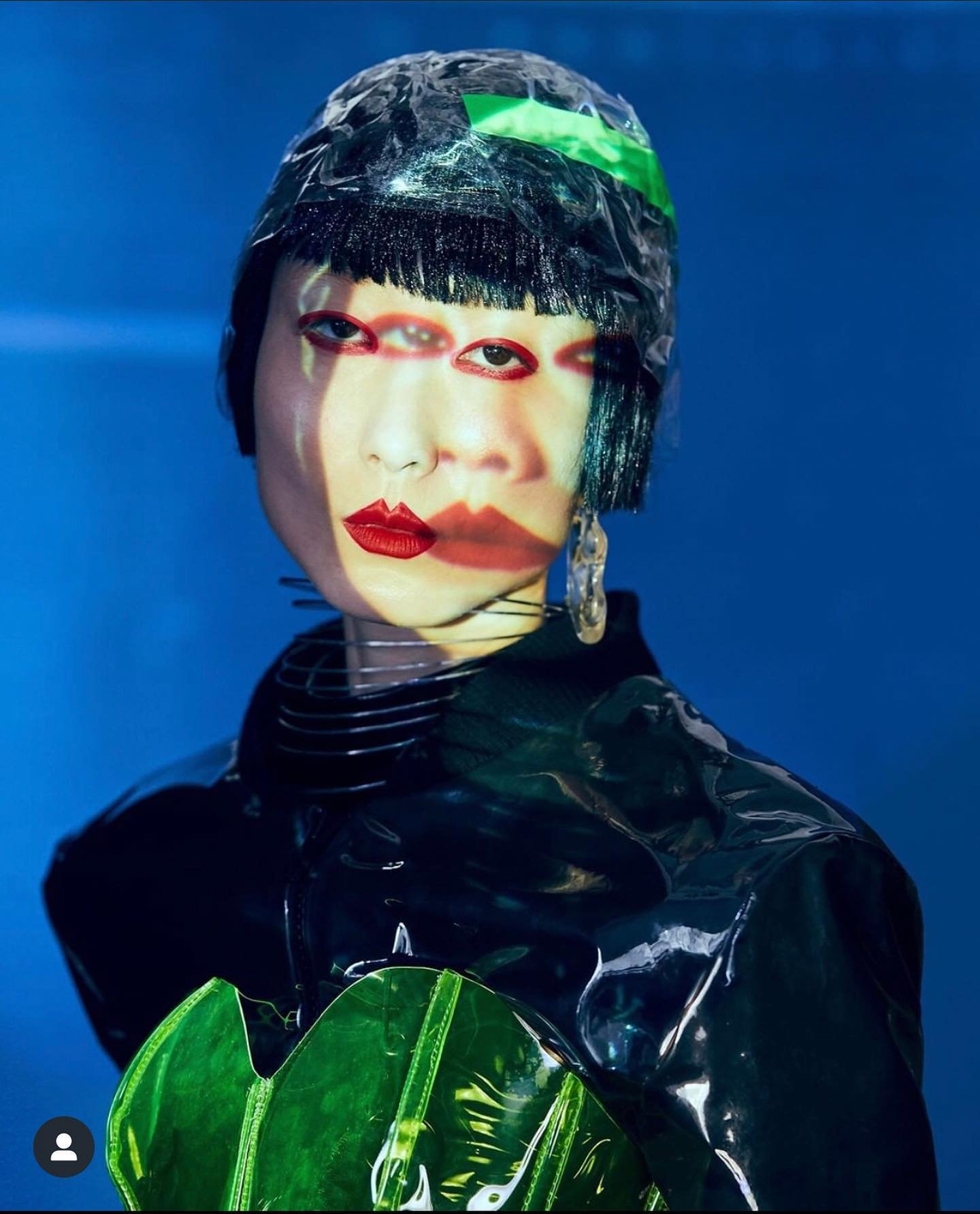 Unique beauty portrait of a person with multiple superimposed facial features in a black glossy garment with futuristic headwear in a blue studio.