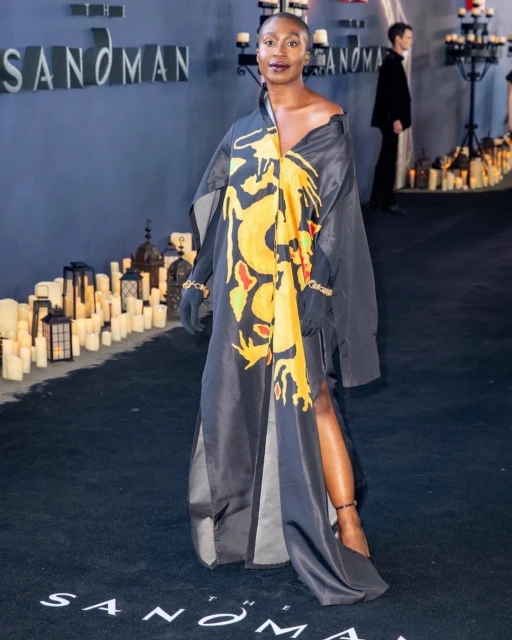 A woman in a black gown with a yellow dragon print poses at 'The Sandman' premiere, with candles and blurred figures in the background.