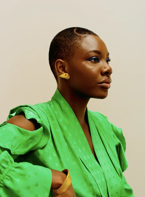 Elegant, dark-skinned woman with short haircut, green dress, gold bangle, and blue eyeliner gazes forward in a studio setting.