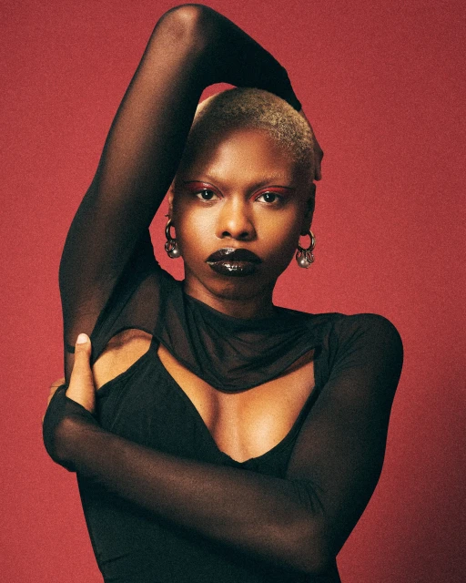 Close-up studio shot of a Black adult female model with a shaved head wearing a sheer black long-sleeved top against a red backdrop.