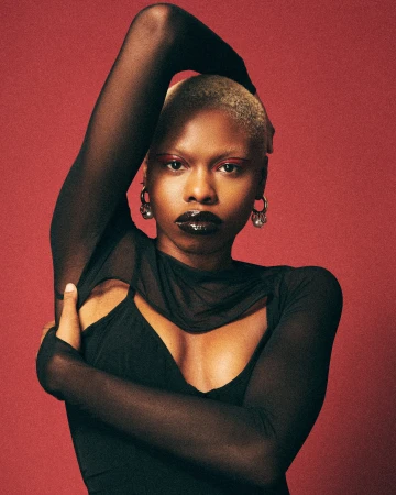 Close-up studio shot of a Black adult female model with a shaved head wearing a sheer black long-sleeved top against a red backdrop.