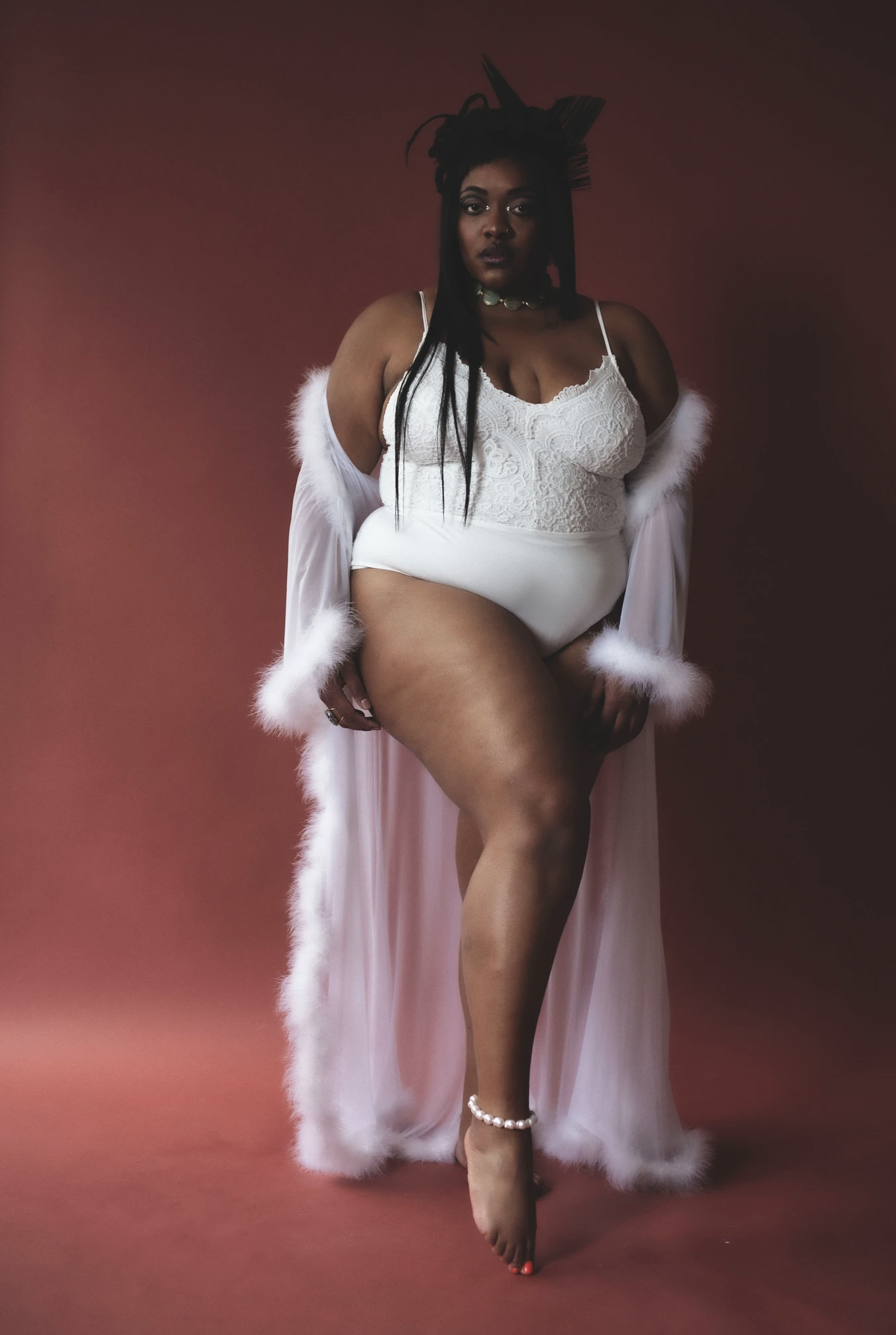 Full body shot of a plus-size adult female in a white lingerie and sheer robe, posing indoors against a terracotta colored background.