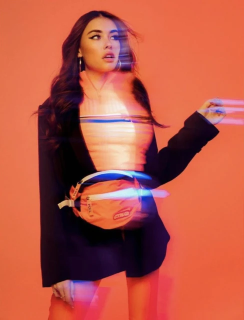 A woman with long dark hair poses indoors wearing a black blazer, light top, and orange fanny pack against an orange background.