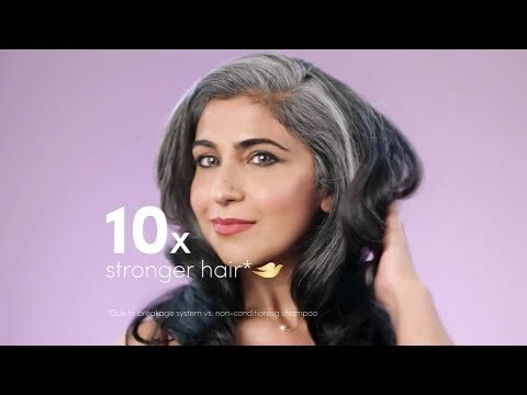 A woman with graying black hair poses in a close-up shot against a light purple backdrop, promoting hair care with visible branding and text.