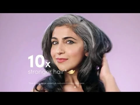 A woman with graying black hair poses in a close-up shot against a light purple backdrop, promoting hair care with visible branding and text.