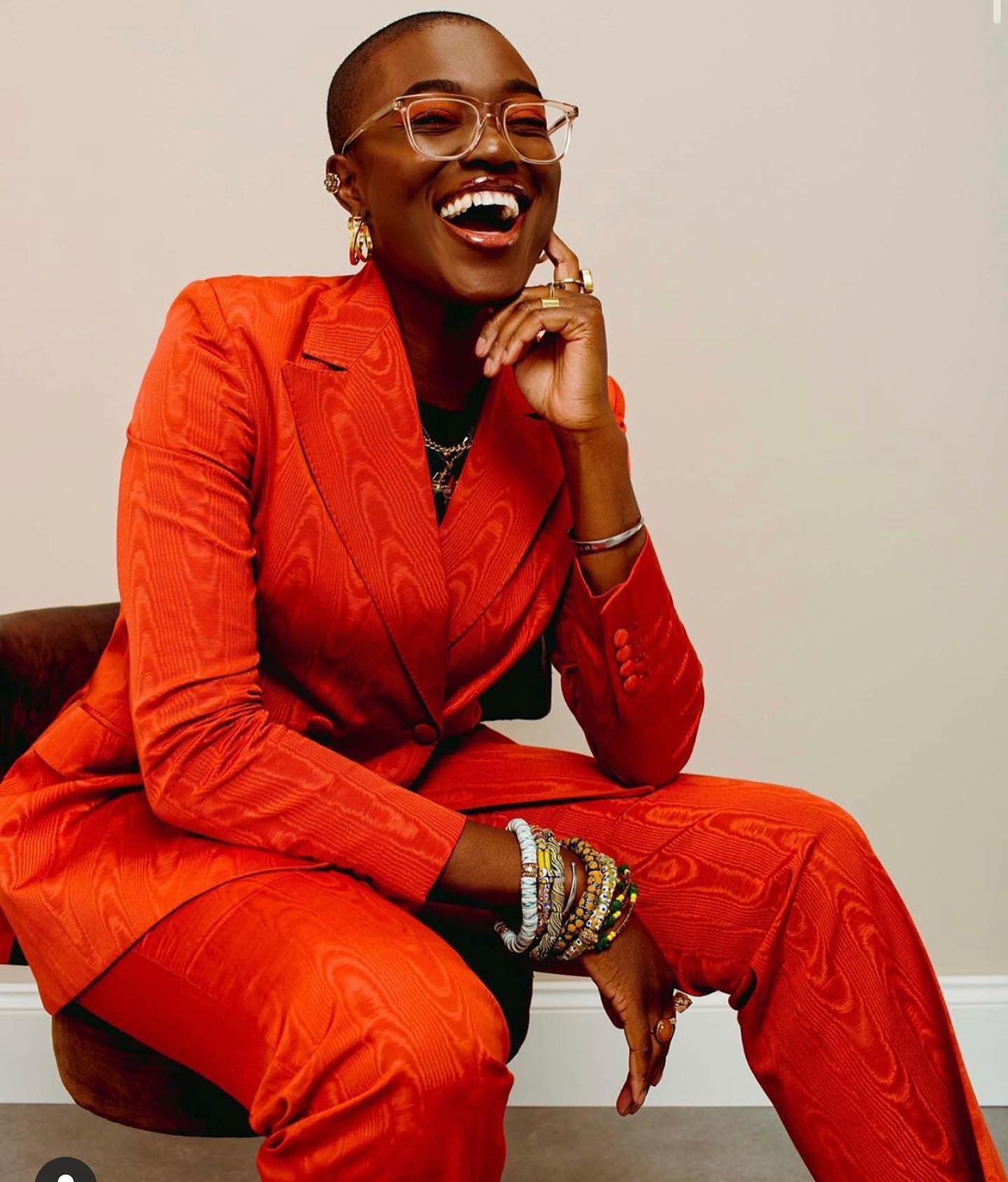 Stylish Black woman in an orange patterned suit, glasses, and jewelry smiles while sitting in a neutral setting, radiating confidence.