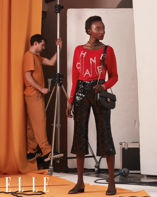 An adult Black woman poses in a red sweater and black sequined pants in a photo studio with a photographer and lighting equipment.