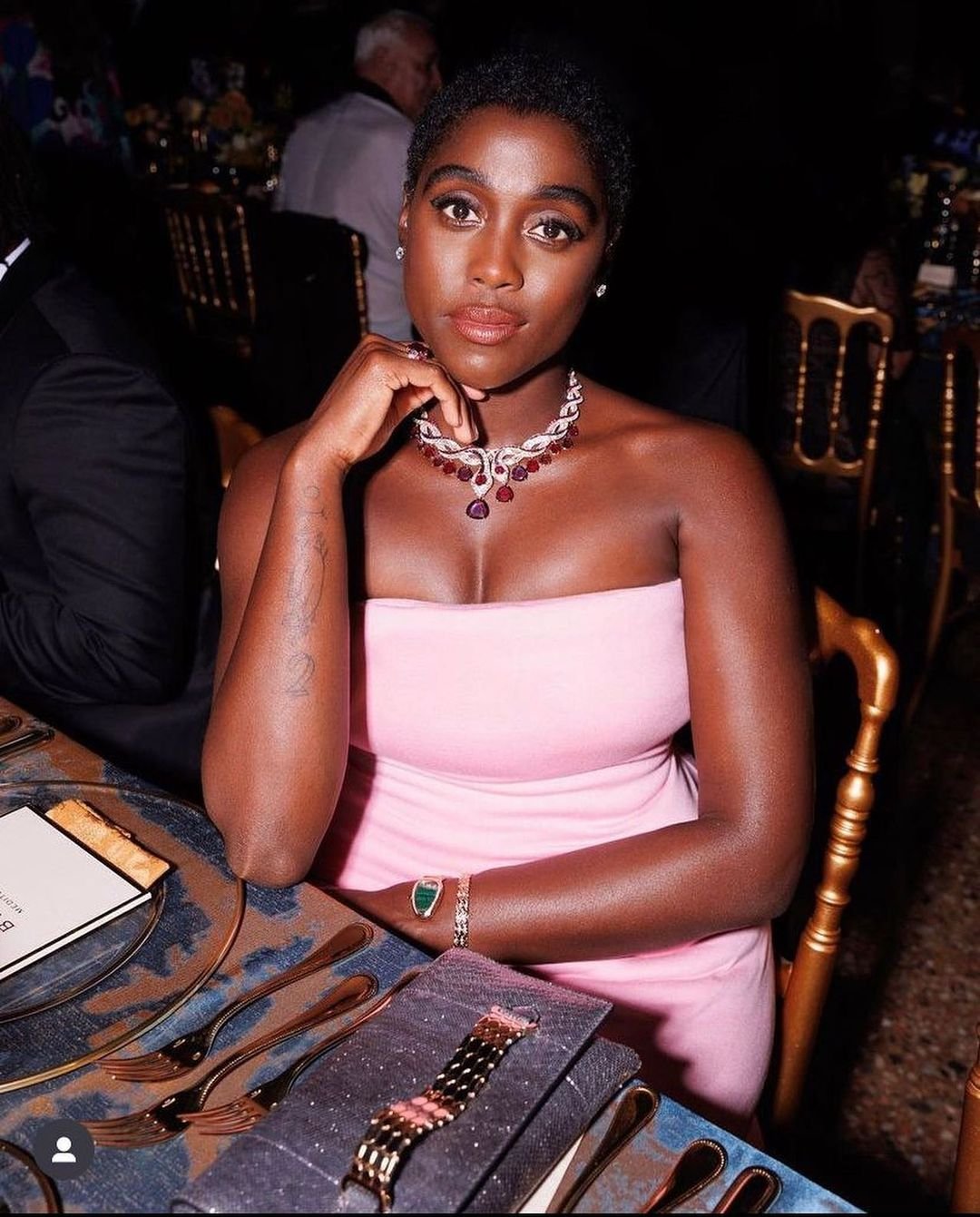 Elegant dark-skinned woman in a pink dress at a formal event, wearing a statement necklace and bracelet, seated at a dining table.