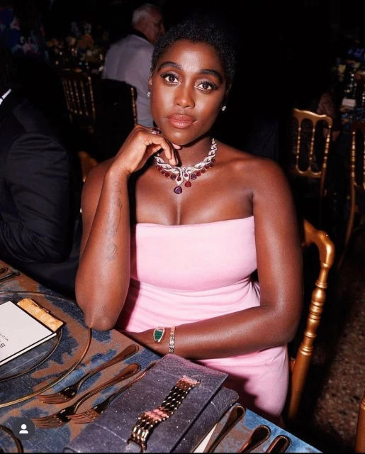 Elegant dark-skinned woman in a pink dress at a formal event, wearing a statement necklace and bracelet, seated at a dining table.