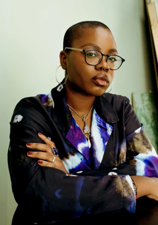 Portrait of a Black woman with short shaved hair wearing glasses and a patterned jacket with arms crossed.