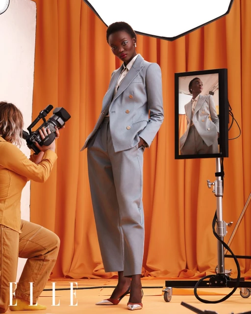 Woman in suit poses as videographer films on orange backdrop in studio with mirror reflecting model for fashion layout.