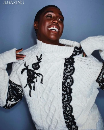 Black woman in a white cable-knit sweater with black lace appliqués laughing against a blue studio background.