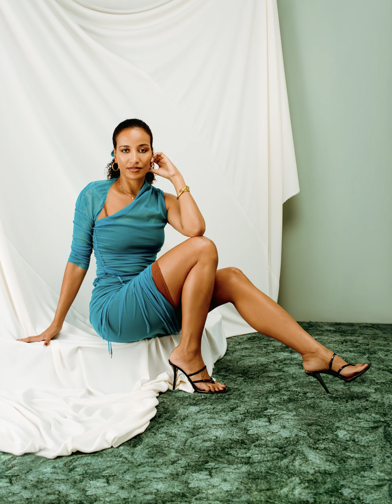 Woman in a teal dress sitting on a green and white backdrop. She wears black heeled sandals and gold jewelry in a studio setting.