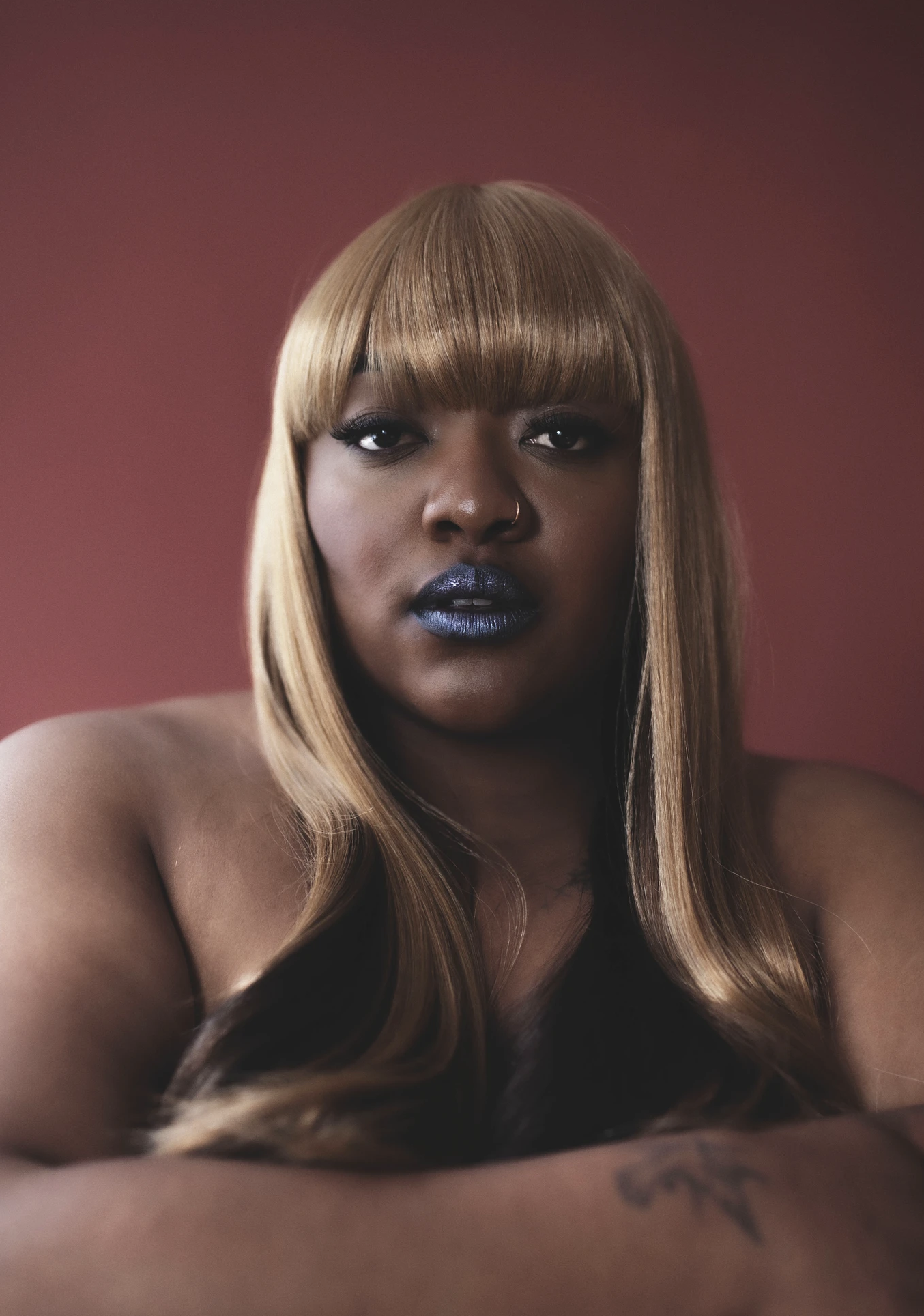 Close-up portrait of an adult female with blonde hair and blue lipstick, posing with her arms crossed against a plain red background.