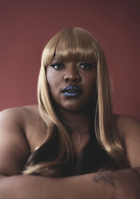Close-up portrait of an adult female with blonde hair and blue lipstick, posing with her arms crossed against a plain red background.