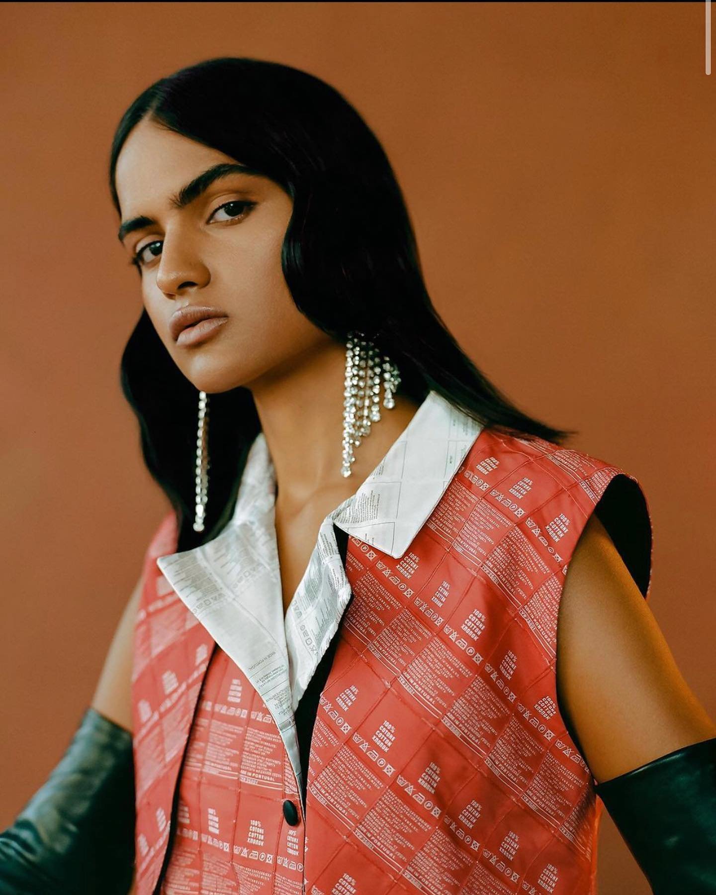 Portrait of a brunette person wearing a red and white vest, long black gloves, and chandelier earrings against a solid brown backdrop.