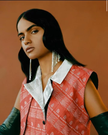 Portrait of a brunette person wearing a red and white vest, long black gloves, and chandelier earrings against a solid brown backdrop.