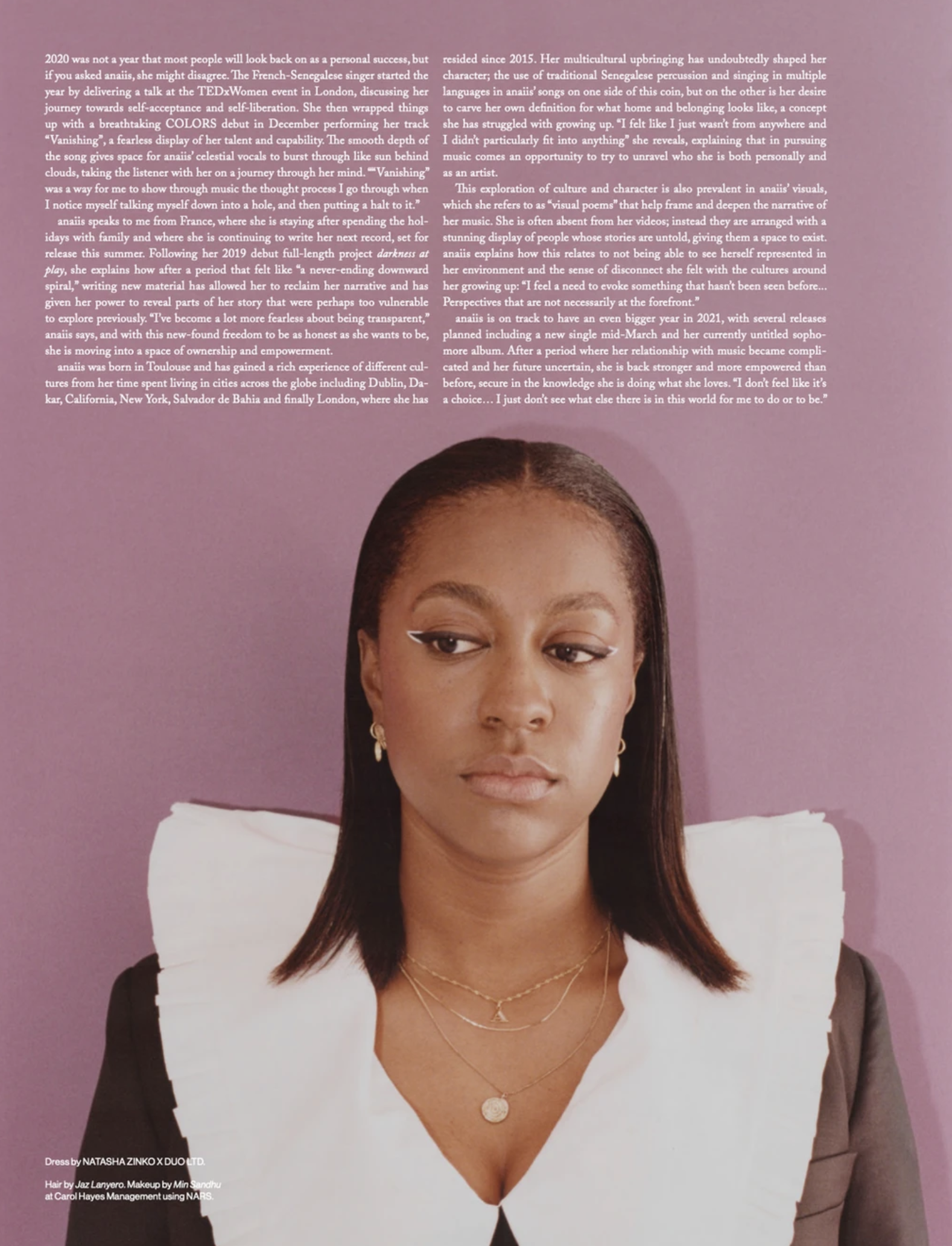 Portrait of a woman in a black blazer with a large white collar, adorned with gold necklaces, against a violet-toned background.