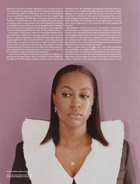 Portrait of a woman in a black blazer with a large white collar, adorned with gold necklaces, against a violet-toned background.