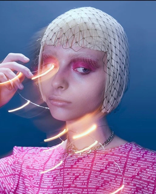 Close-up of a young adult woman with pink eye makeup, a net head covering, and a pink patterned shirt against a blue background.