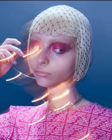 Close-up of a young adult woman with pink eye makeup, a net head covering, and a pink patterned shirt against a blue background.