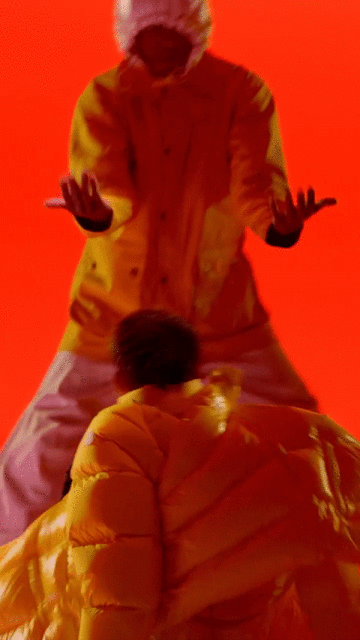 Two people in yellow puffy coats are set against a bright orange backdrop, creating a vibrant and eye-catching vertical studio shot.