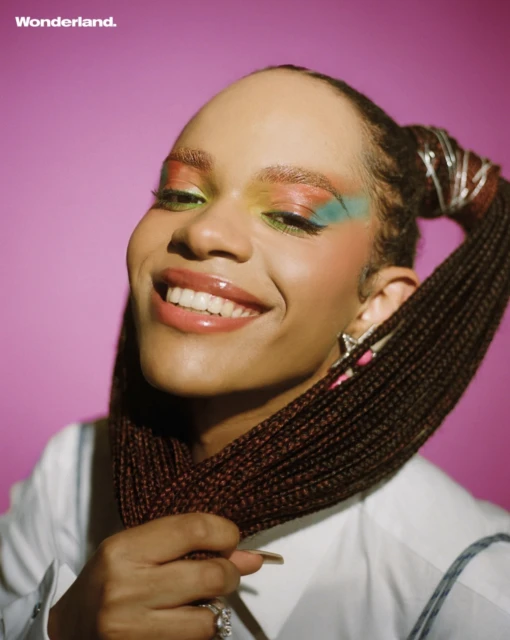 Close-up portrait of a smiling woman with box braids, colorful eyeshadow, and a white shirt against a pink background, editorial fashion.