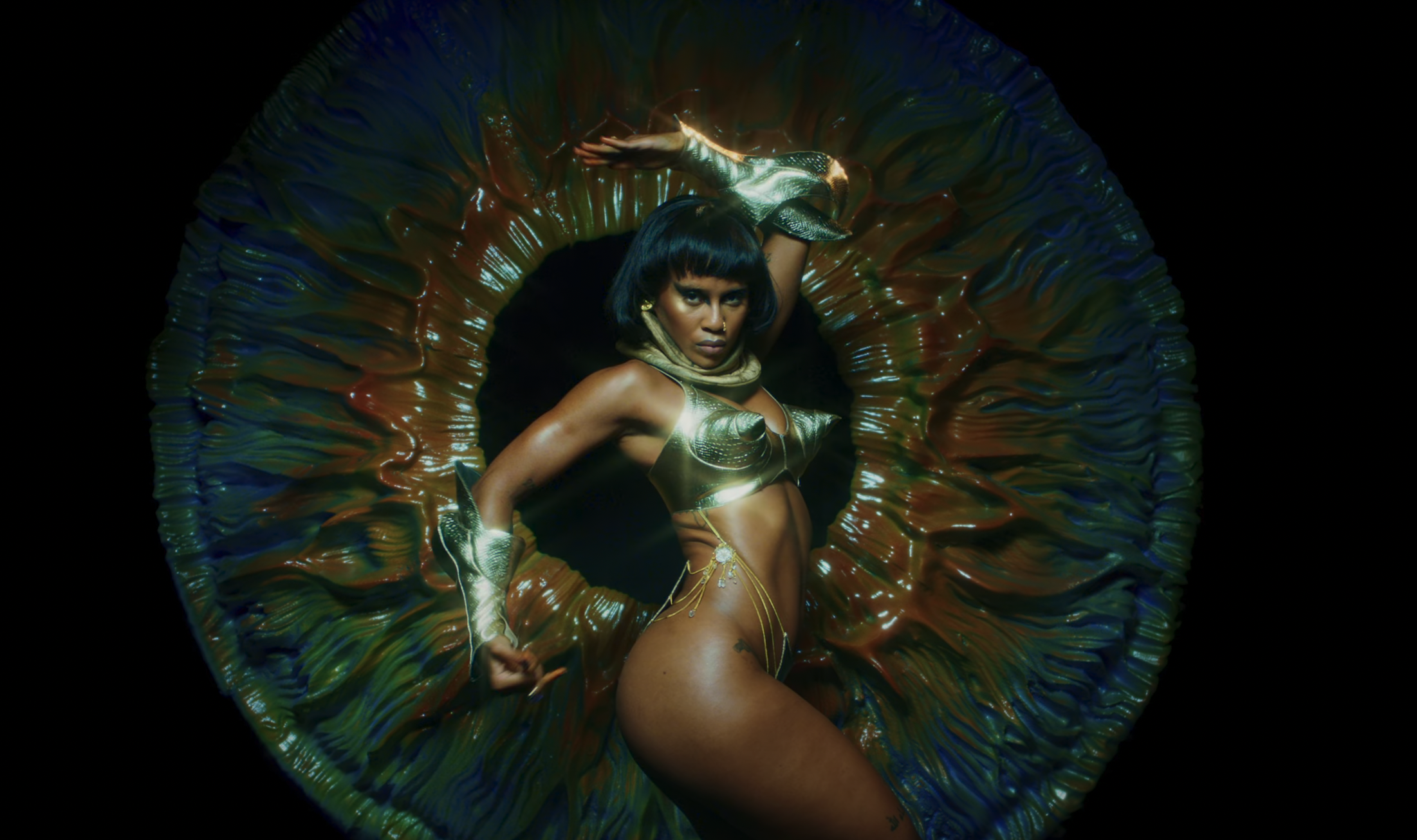 Adult woman in gold attire poses dynamically in front of an abstract, organic-looking floral sculpture against a black backdrop.