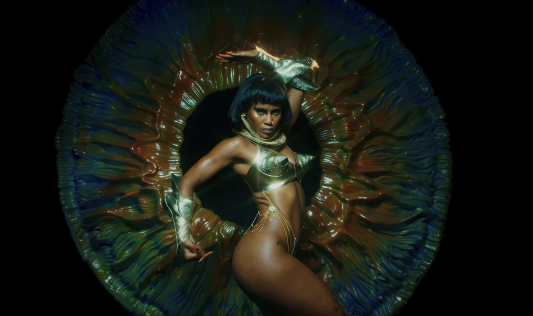 Adult woman in gold attire poses dynamically in front of an abstract, organic-looking floral sculpture against a black backdrop.