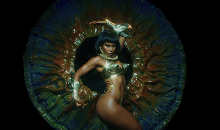 Adult woman in gold attire poses dynamically in front of an abstract, organic-looking floral sculpture against a black backdrop.