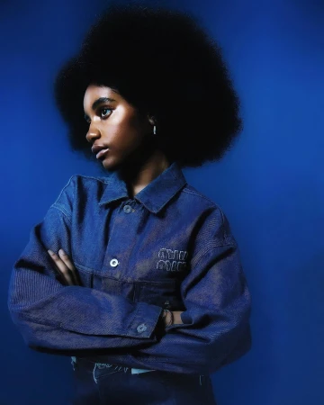 Portrait of a Black woman with a large afro wearing a denim jacket against a solid blue background.