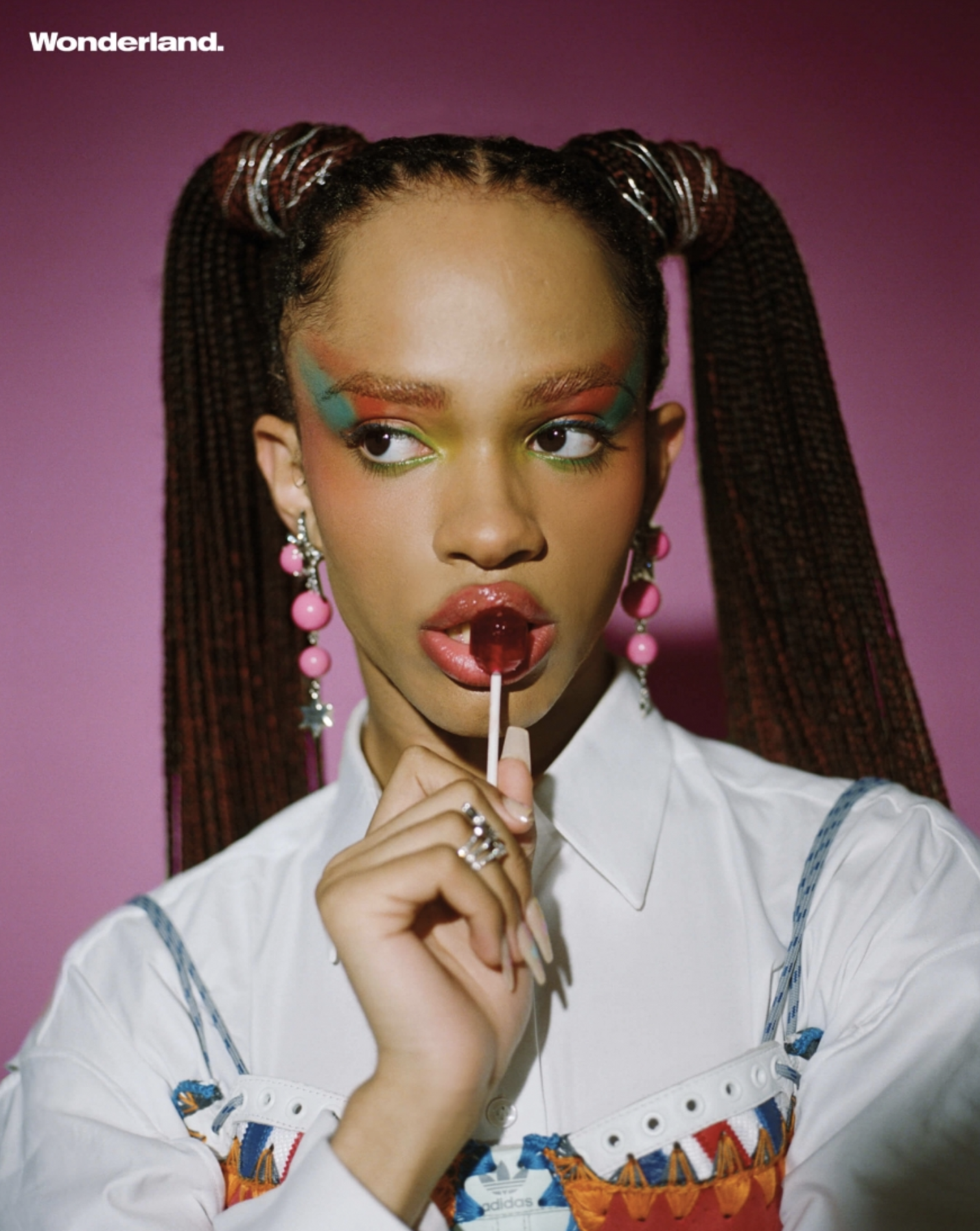 Close-up portrait of a fashionable young woman with rainbow makeup, braided pigtails, jewelry, a white shirt, and a pink lollipop.