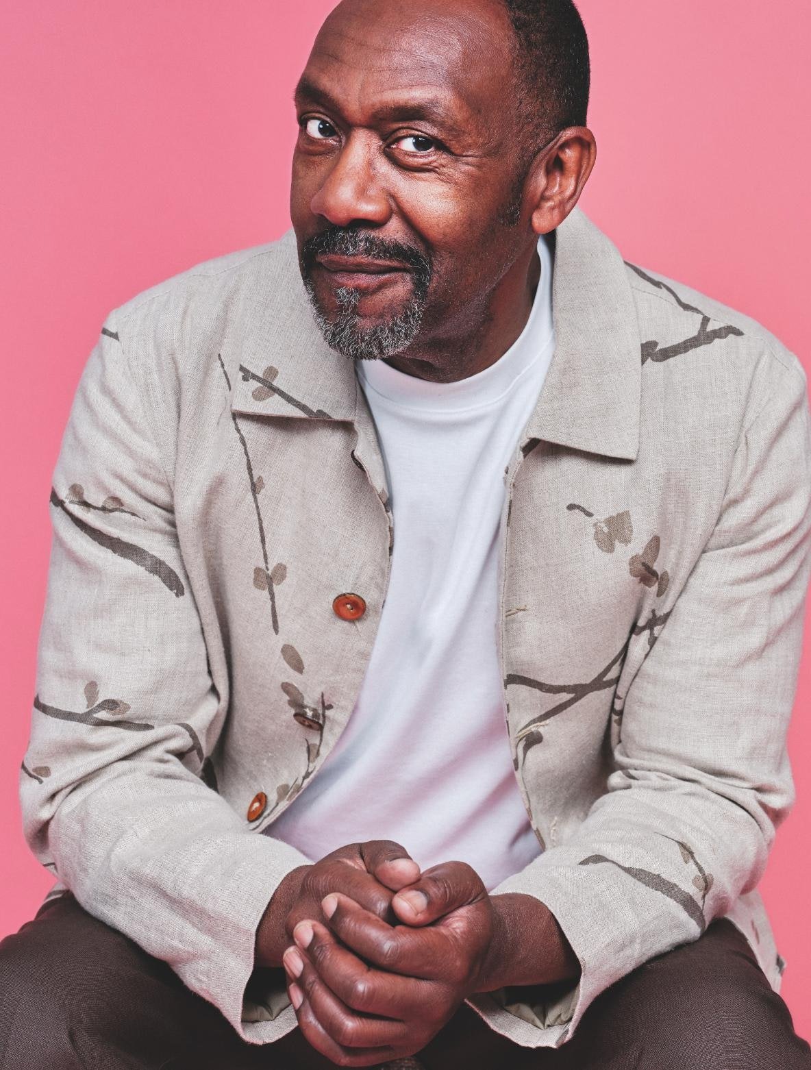 Portrait of a handsome Black man with a salt-and-pepper beard wearing a patterned jacket and T-shirt against a pink background.