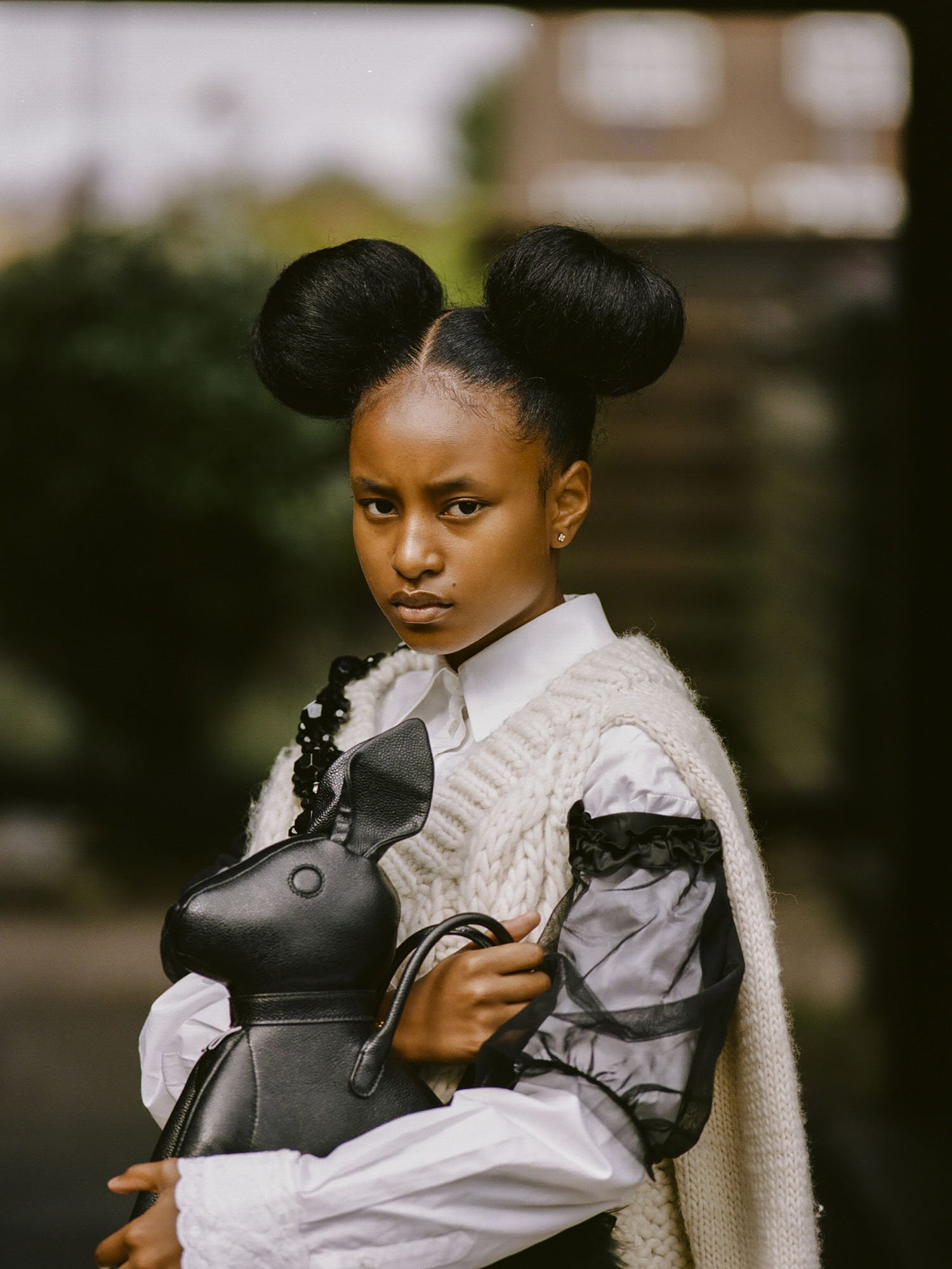 Portrait of a young Black female wearing a white shirt, grey knit, and unique hairstyle, holding a black bunny-shaped bag in a blurred setting.