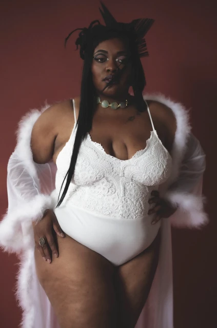 Plus-size African American woman in a white bodysuit and sheer robe poses against a reddish-brown background in a studio setting.
