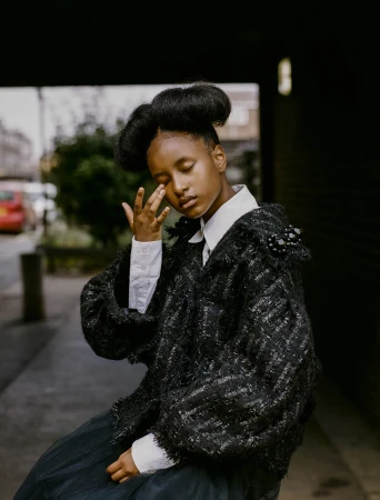 A young person with styled hair wearing a collared shirt and black jacket poses outdoors, looking downward with hand near their face.
