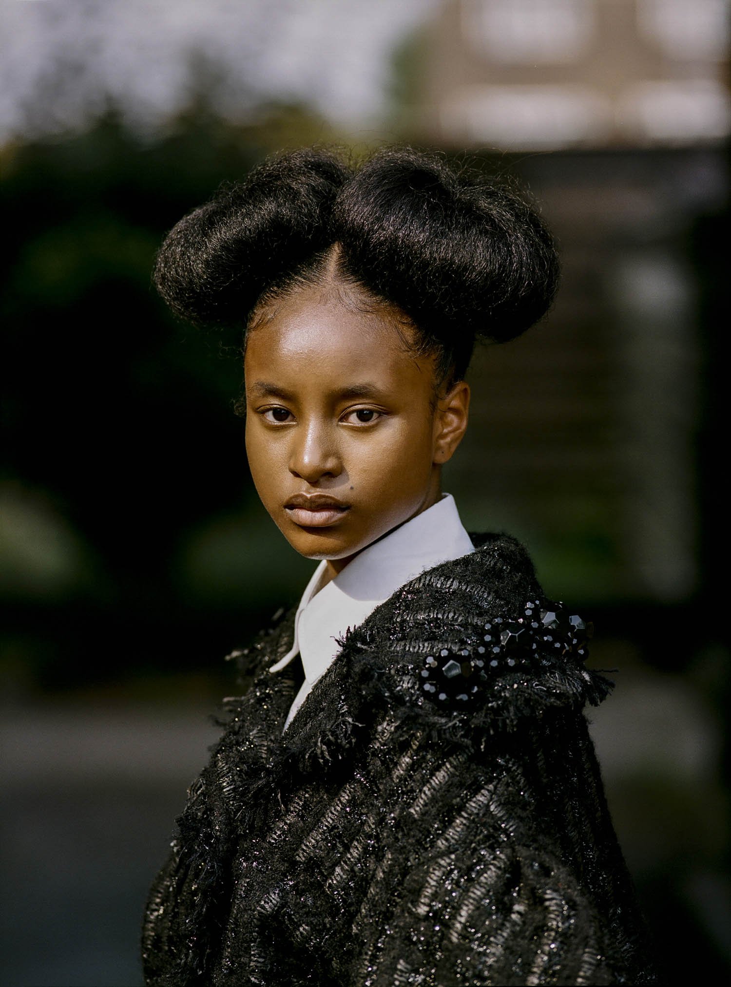 Portrait of a young black woman with an elaborate updo, wearing a textured black jacket with embellishments and a white collared shirt. 