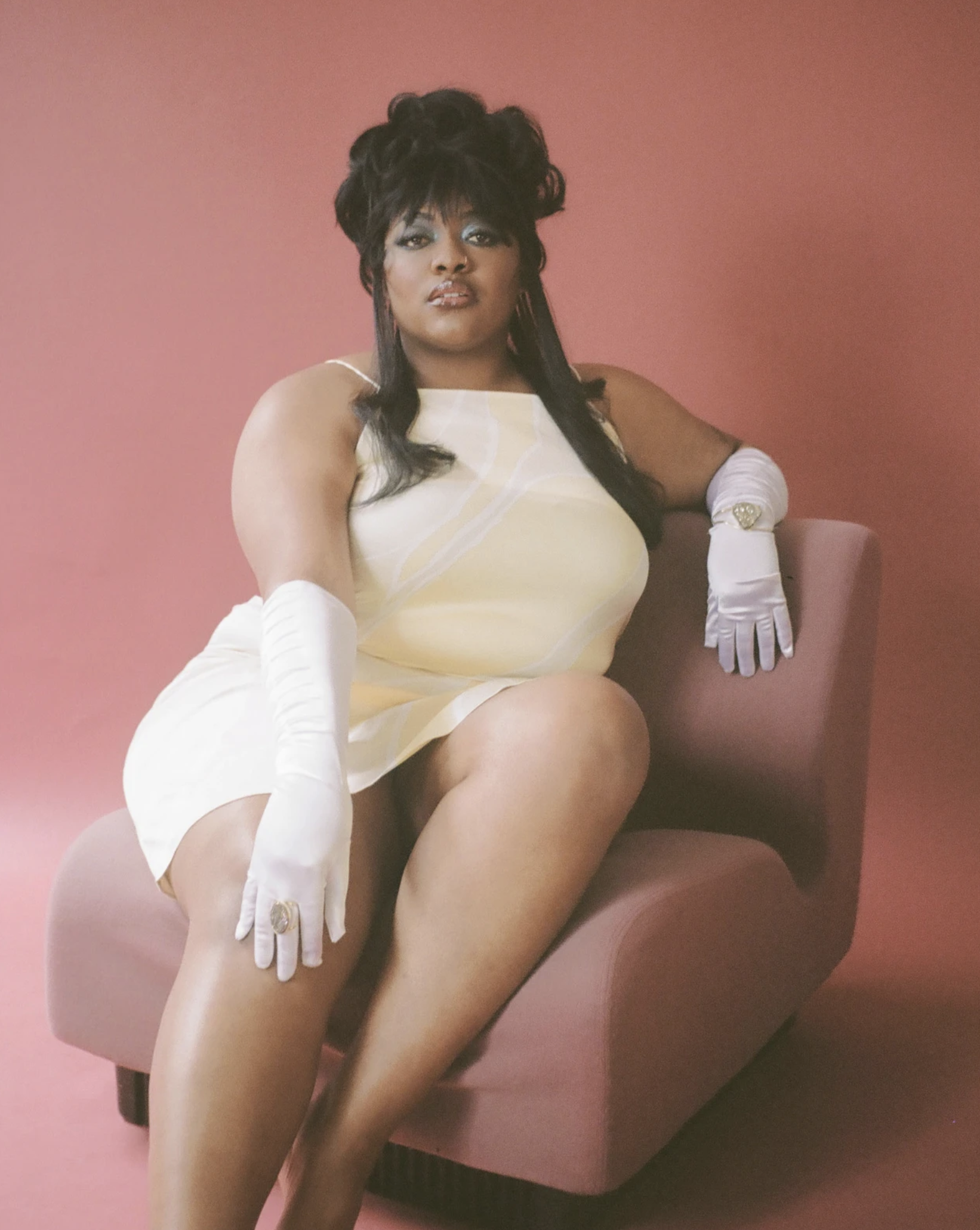 A woman with dark hair in a yellow dress and white gloves sits in a mauve chair against a salmon-pink backdrop in a studio shot.