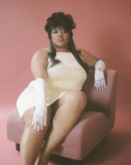 A woman with dark hair in a yellow dress and white gloves sits in a mauve chair against a salmon-pink backdrop in a studio shot.