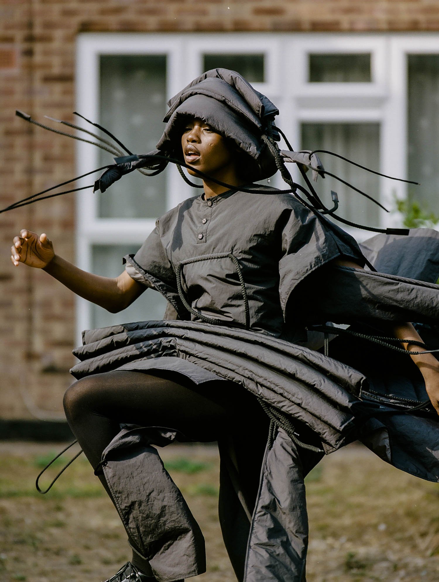 A Black female fashion model in an avant-garde gray quilted outfit poses outdoors on a grassy lawn in front of a brick house.