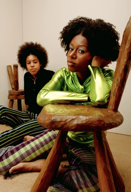Two Black women in an indoor studio setting, one seated on the floor and one sitting on a carved wooden chair in vibrant green attire.