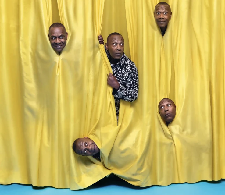 African-American man in a printed shirt stands behind a bright yellow curtain with 4 headshots popping through holes in the fabric.