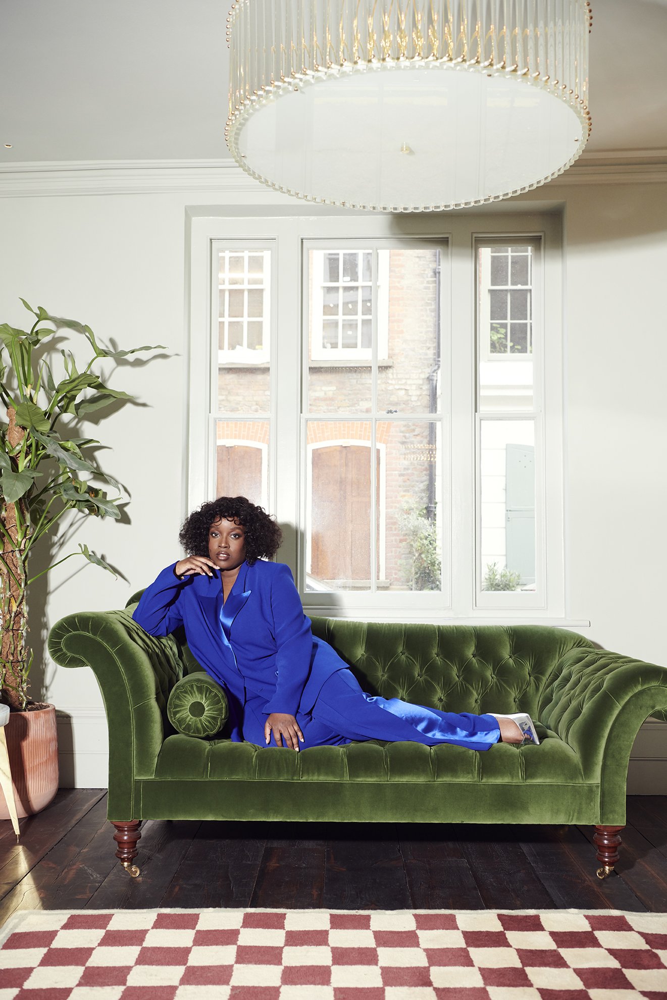 An African American woman poses on a green velvet couch in a blue suit, inside a white-walled room with a window and patterned carpet.
