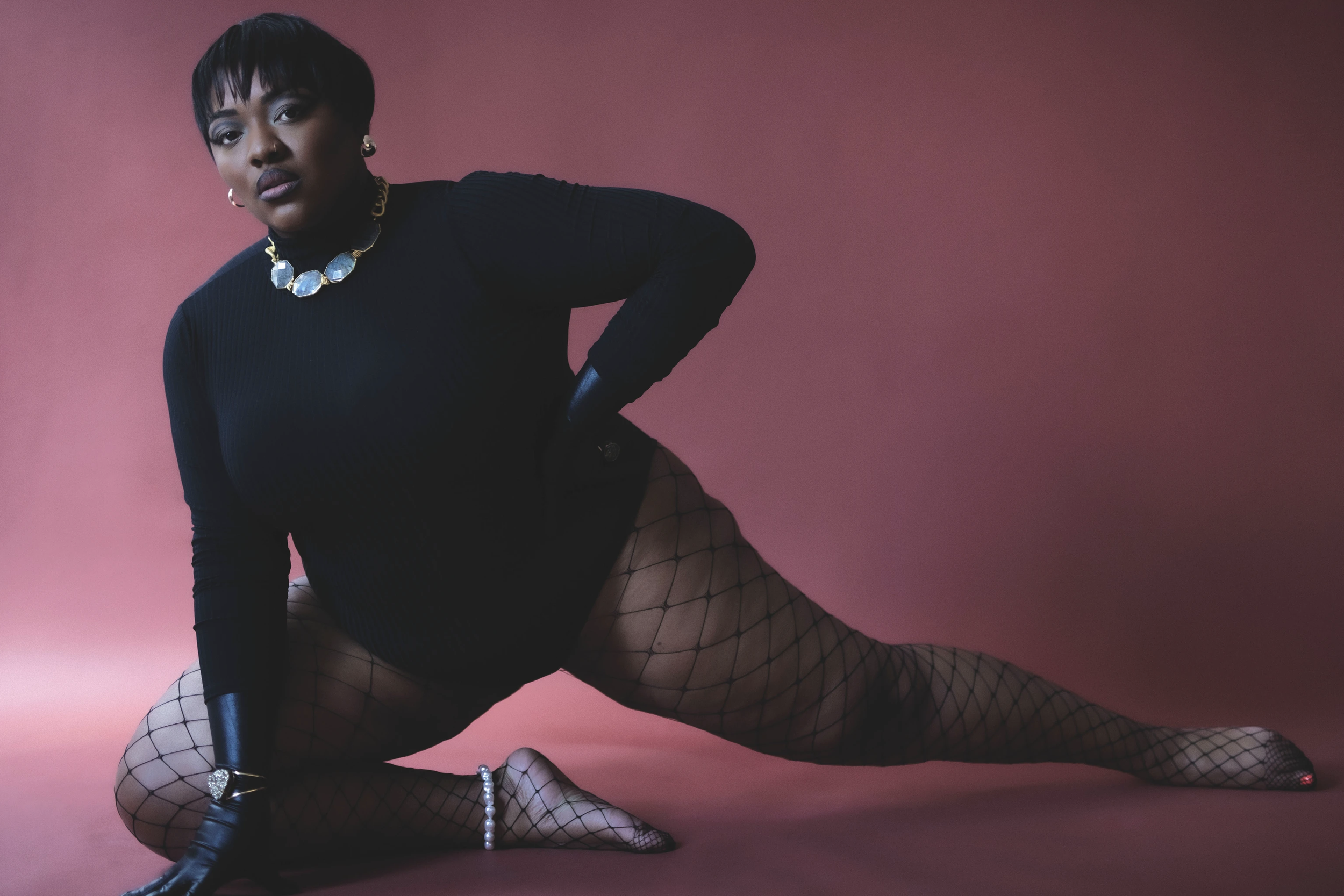 An adult female model in black bodysuit and fishnet tights strikes a dynamic pose against a dark-red background. She accessorizes with jewelry.