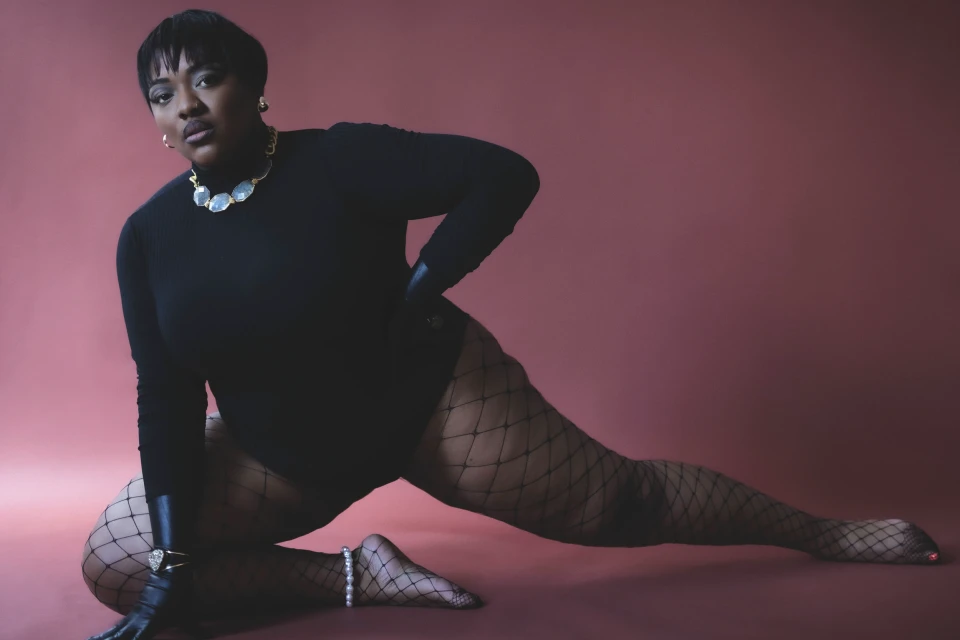 An adult female model in black bodysuit and fishnet tights strikes a dynamic pose against a dark-red background. She accessorizes with jewelry.