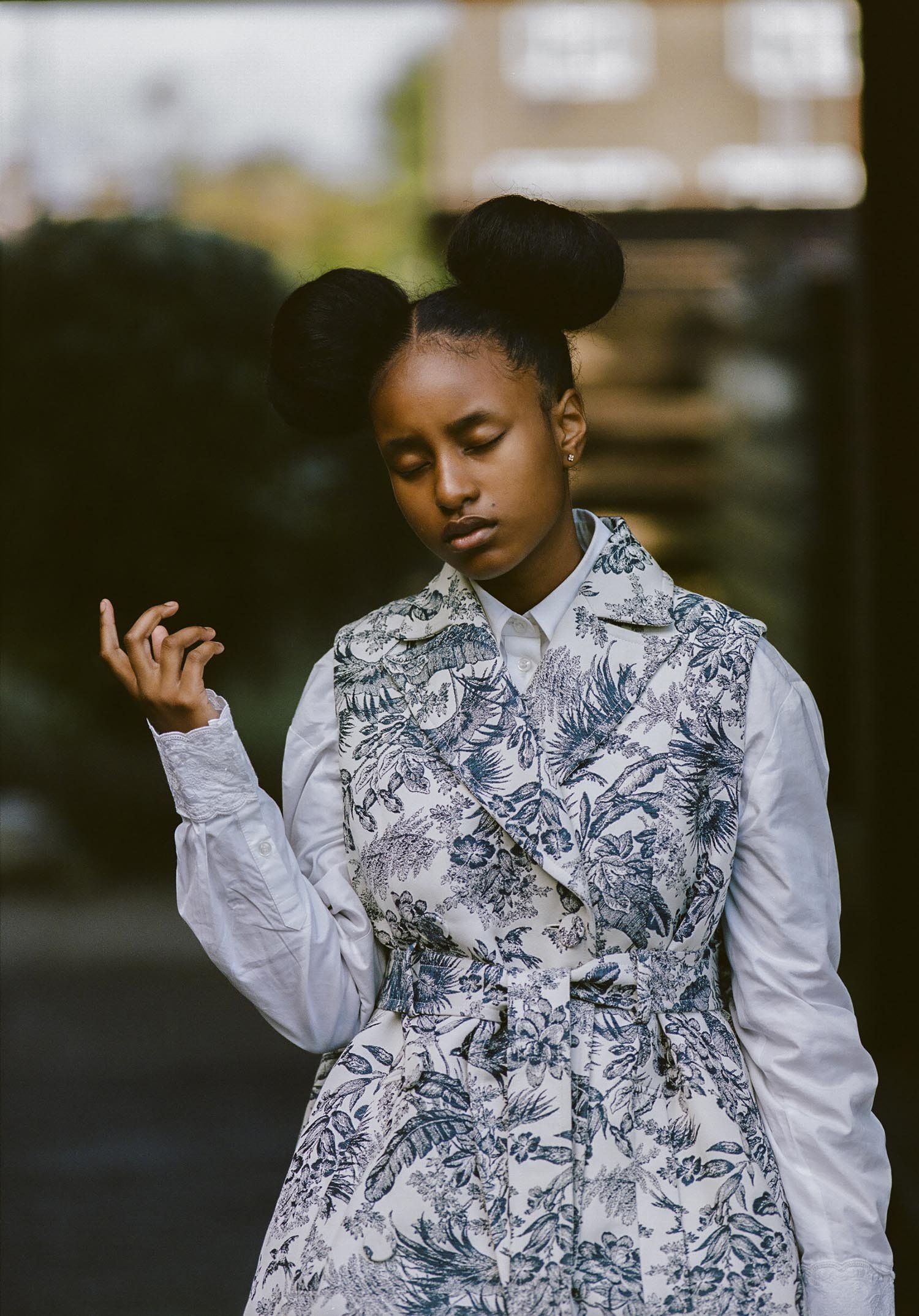 Young woman in an elegant patterned vest and blouse posing outdoors with a serene expression and space buns hairstyle.